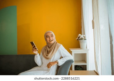 portrait of Asian veiled woman casually sitting on sofa using cell phone at home - Powered by Shutterstock