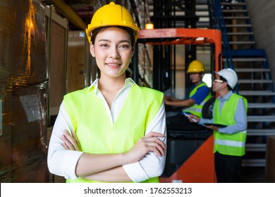 Portrait of Asian warehouse woman worker smile with arms crossed, supervisor talking with forklift driver at the background. export import or logistics service business. employee and labor factory - Powered by Shutterstock