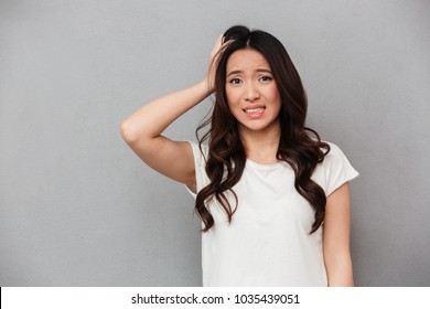 Portrait of asian woman 20s with dark curly hair touching her head and expressing worry isolated over gray background