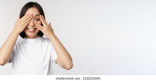 Portrait Of Asian Woman Covering Eyes, Waiting For Surprise Blindfolded, Smiling And Peeking At Camera, Standing Over White Background