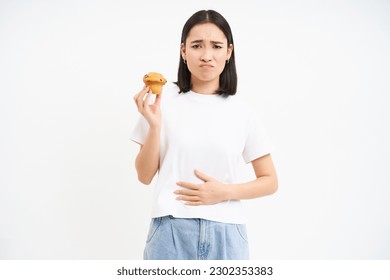 Portrait of asian woman feeling pain in stomach, discomfort after food, holding cupcake and looking unhappy, white background. - Powered by Shutterstock