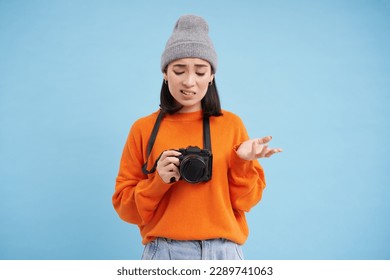 Portrait of asian woman in hat, holding digital camera with confused face, unprofessional photographer doesnt know how to take pictures on digicam, blue background.