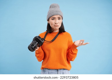Portrait of asian woman in hat, holding digital camera with confused face, unprofessional photographer doesnt know how to take pictures on digicam, blue background.