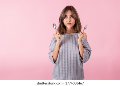 Portrait Of An Asian Woman Holding A Spoon And Fork In His Hand. She Is Choosing A Food Menu. Shot Of The Studio. 
Separately On The Pink Background.