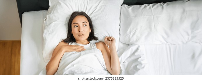 Portrait of asian woman lying in bed with shocked face, looking startled and upset, gasping from smth, staying alone in her bedroom.