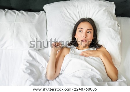 Portrait of asian woman lying in bed with shocked face, looking startled and upset, gasping from smth, staying alone in her bedroom. Stockfoto © 