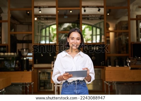 Portrait of asian woman, manager standing with tablet in front of cafe entrance, welcomes guests. Business and entrepreneurs concept