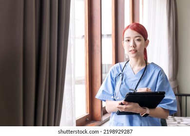 Portrait Of Asian Woman Nurse Wearing Medical Scrubs, Stethoscope And Holding The Tablet In The House Of Patient. Caregiver Visit At Home. Home Health Care And Nursing Home Concept.