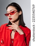 portrait of asian woman in red stylish sunglasses and ear cuff touching chin isolated on grey
