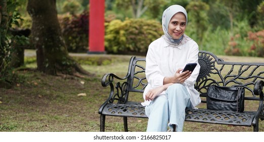 Portrait Of Asian Woman Sitting In The Park Bench Holding  Cell Phone