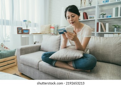 Portrait Asian Woman Sitting With Pillow On The Sofa In Casual Wear Is Using Her Credit Card To Pay While Shopping Online On Phone In A Bright Home Living Room.