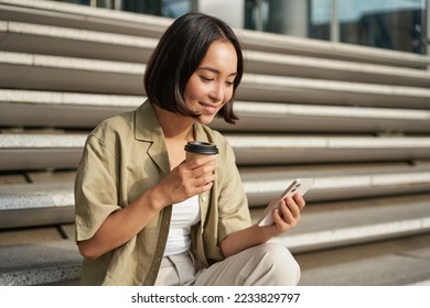 Portrait of asian woman with smartphone, drinks coffee and watches videos on mobile phone. Girl with telephone sits on stairs outdoors. - Shutterstock ID 2233829797