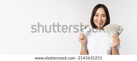 Portrait of asian woman smiling, holding credit card and money cash, dollars, standing in tshirt over white background