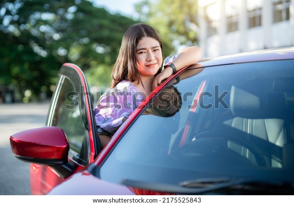Portrait asian\
woman Wear purple dress wearing smartwatch in the car door modern\
red car at the city park\
outdoors.