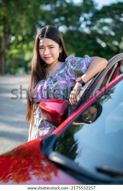 Portrait asian\
woman Wear purple dress wearing smartwatch in the car door modern\
red car at the city park\
outdoors.