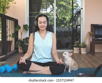 Portrait of Asian woman wearing headphones, practice meditation on yoga mat in balcony  with computer laptop  and Chihuahua dog. Yoga or meditation training online.