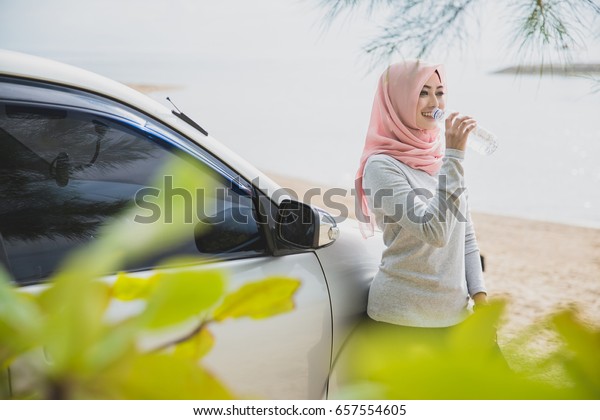 portrait of asian woman wearing hijab drinking\
mineral water while lying on the\
car