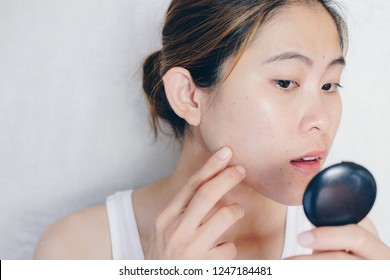 Portrait Of Asian Woman Worry About Her Face When She Saw The Problem Of Acne And Scar By The Mini Mirror. Conceptual Shot Of Acne & Problem Skin On Female Face.