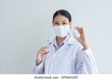 Portrait of Asian young doctor or scientist woman holding showing Covid-19 vaccines vial bottle in hand with face mask, white gown and holding syringe in other hand. White background.  - Shutterstock ID 1993641587