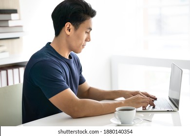 Portrait Of Asian Young Man Sit Near Bookshelf Using Notebook Laptop. Handsome Guy Wear Dark Blue Shirt Typing On Computer Keyboard. White Coffee Cup On Work Desk. Copy Space.