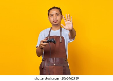 Portrait Of Asian Young Man Wearing Apron Holding Game Controller And Making Stop Motion With Hands Over Yellow Background