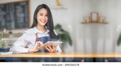 Portrait Of Asian Young Woman Cafe Owner With The Tablet, Image Of An Asian Woman Barista Wearing An Apron At The Café.