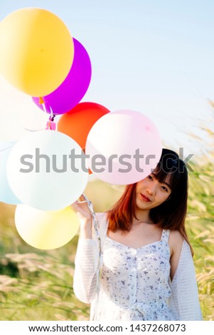 Portrait of asian young woman with smiling face, holding a bunch of colorful balloons and walking on grassland during sunset background and flair light