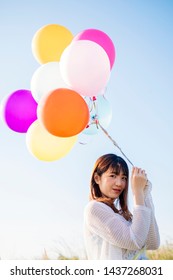 Portrait of asian young woman wearing white dress holding a bunch of colorful balloons and smilling against blue sky background 
