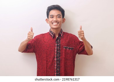 Portrait Of The Asian Youth Smiling Broadly And Holding Up His Two Thumbs In A Red Batik Shirt. Isolated Image On A White Background 