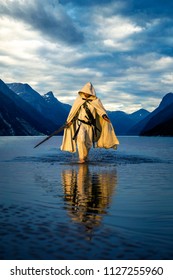 Portrait of assassin in white costume with the sword at the sea. He is posing near water with reflections on it. Sunset time. Isolated.