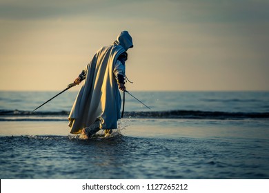 Portrait of assassin in white costume with the sword at the sea. He is posing near water during sunset, soft light.