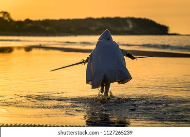 Portrait of assassin in white costume with the sword at the sea. He is posing near water during sunset, soft light.