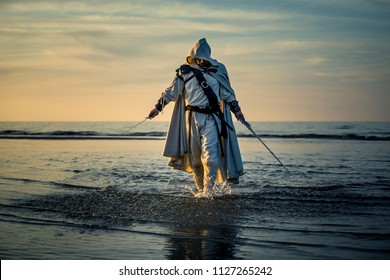 Portrait des Mörders in weißem Kostüm mit dem Schwert am Meer. Er posiert bei Sonnenuntergang nahe Wasser, weiches Licht.