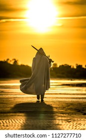 Portrait des Mörders in weißem Kostüm mit dem Schwert am Meer. Er posiert bei schönem Sonnenuntergang am Meer. Gelbes, weiches Licht.