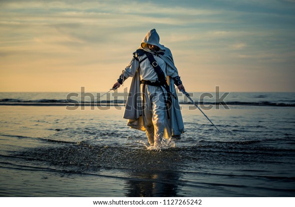 Portrait of\
assassin in white costume with the sword at the sea. He is posing\
near water during sunset, soft\
light.