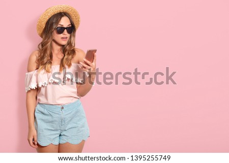 Portrait of astonished pretty woman wearing blouse with bared shoulders, short, straw hat and sunglasses, holds smart phone with surprised facial expression, checking social network, has shocking news