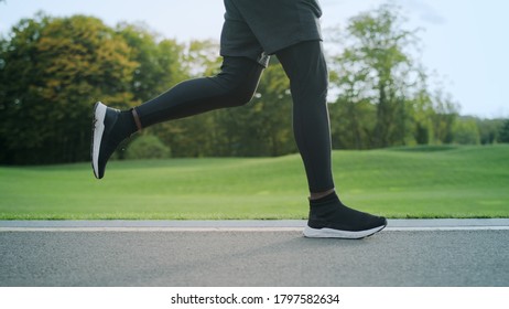 Portrait Of Athletic Afro Man Running In Park. Male Runner Training On Asphalt Surface In Slow Motion. Energetic Man Legs Jogging On Road At Morning