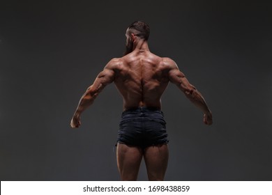 Portrait Of A  Athletic Man With A Naked Torso Showing Muscles Isolated On A Dark Gray Background, View From The Back