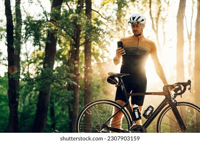 Portrait of an athletic man in sport clothes wearing helmet and glasses and using smartphone while standing near bike outdoors. Concept of people, technology and recreation. Sport cycling.