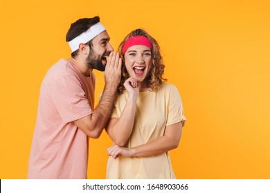 Portrait of athletic young couple wearing headbands wondering while whispering secret or gossip isolated over yellow background
