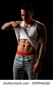 Portrait of athletic young tattooed guy in white undershirt and blue jeans. Black background. Studio shot