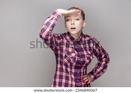 Portrait of attentive concentrated attractive woman with bun hairstyle looking far in distance with hand near forehead, wearing checkered shirt. Indoor studio shot isolated on gray background.