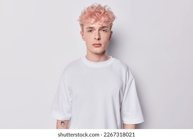 Portrait of attentive hipster guy with pink hair and tattoo on arm dressed in casual basic t shirt looks directly at camera has serious expression isolated over white background belong to subculture