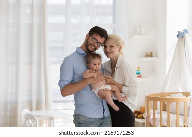 Portrait Of Attractive 30s Parents Holding On Arms Their Baby In Diaper, Smiling Look At Camera Standing In Cozy Nursery Near Modern Crib. Bank Loan For Young Family, Happy Parenthood, Love Concept