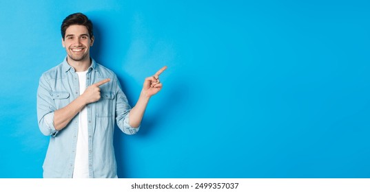 Portrait of attractive adult man smiling, pointing fingers right at logo or banner, showing advertisement against blue background. - Powered by Shutterstock