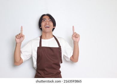 Portrait Of Attractive Asian Barista Man In Brown Apron Showing Product, Pointing Up At Something And Smiling. Advertising Concept. Isolated Image On White Background