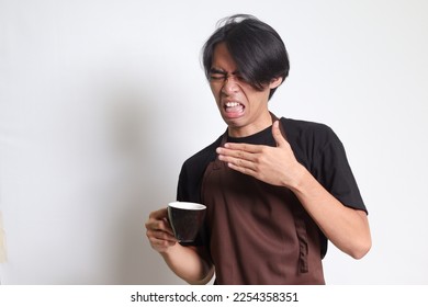 Portrait of attractive Asian barista man in brown apron making unpleasant face while drinking a cup of coffee. Isolated image on white background