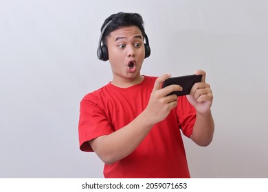 Portrait of attractive Asian man in red t-shirt using wireless headphone playing games on his mobile phone by tilting the screen. Wow face expression. Isolated image on gray background