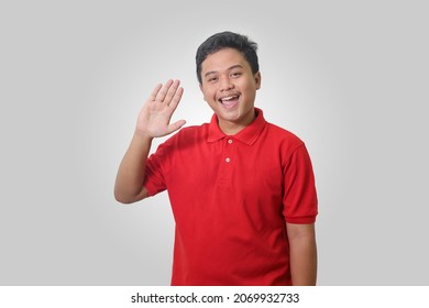 Portrait Of Attractive Asian Man In Red Polo Shirt Saying Hi, Waving Hand At Camera, Greeting Person With Happy Smile. Isolated Image On White Background