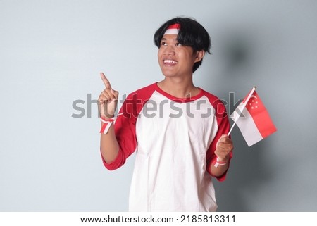 Portrait of attractive Asian man in t-shirt with red and white ribbon on head, showing product and poiting away while holding singapore flag. Isolated image on gray background